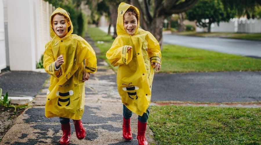 Two children in yellow bee raincoats and red boots playing outside on wet pavement, showcasing carefree childhood joy.